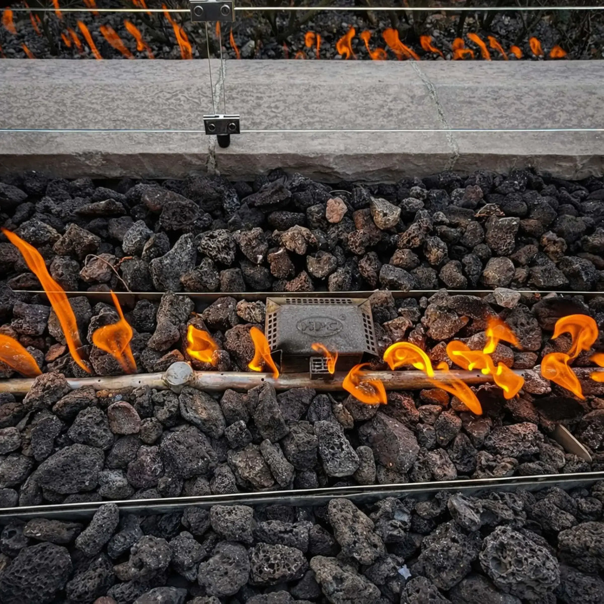 Rustic metal fire bowl with a tall, dramatic flame rising from lava rock, placed in a landscaped yard with flowering plants, stone edging, and a wooden staircase in the background.