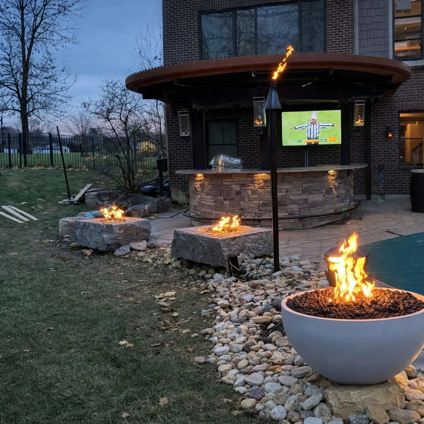 Modern black rectangular fire table filled with lava rock and low flames, surrounded by metal mesh patio chairs and café tables on a paved outdoor dining area with a yellow umbrella and street view beyond.