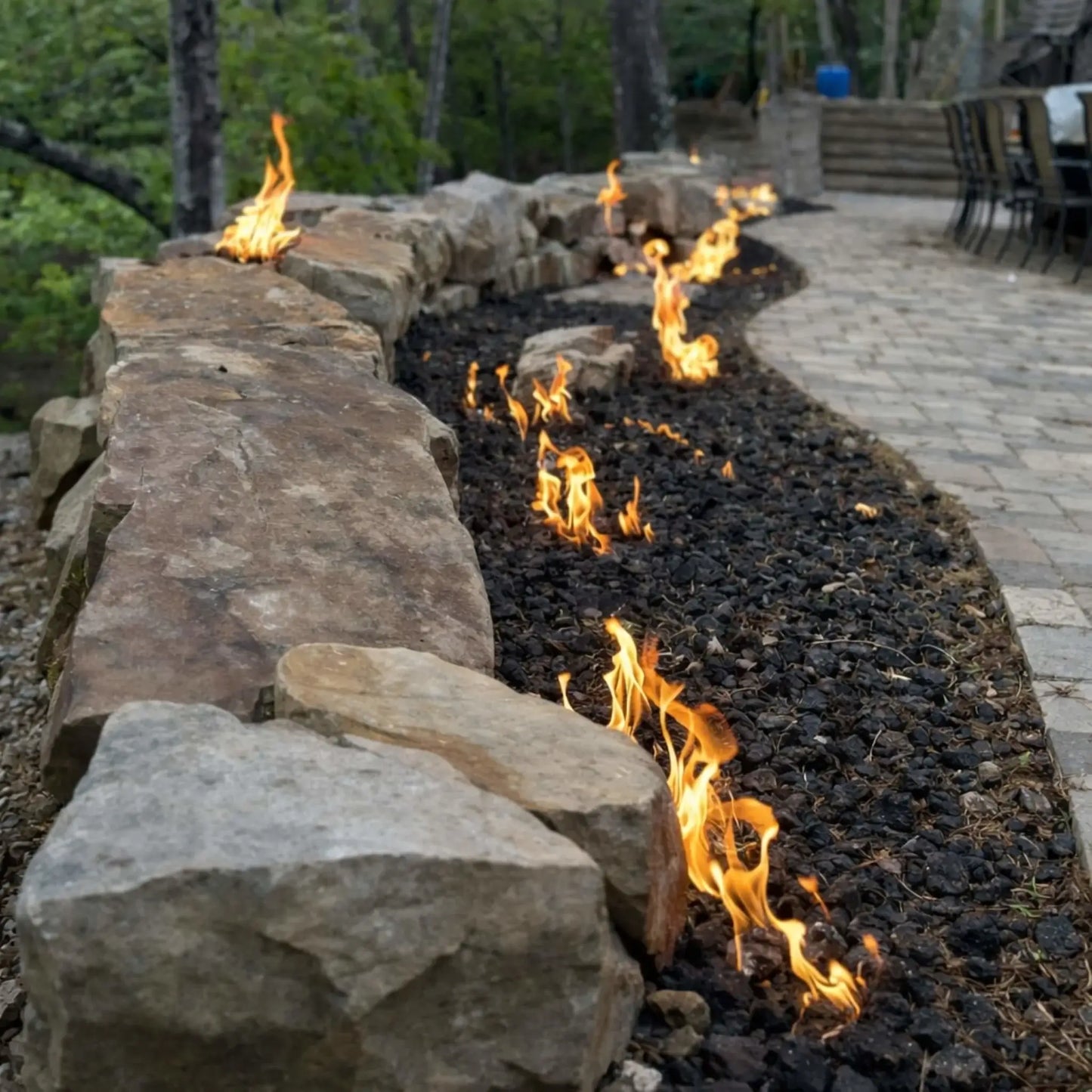 Curved outdoor gas fire feature built into a natural stone retaining wall, with multiple flame points rising through black lava rock along a landscaped patio edge, surrounded by trees and a paved walkway for a warm, upscale backyard design.