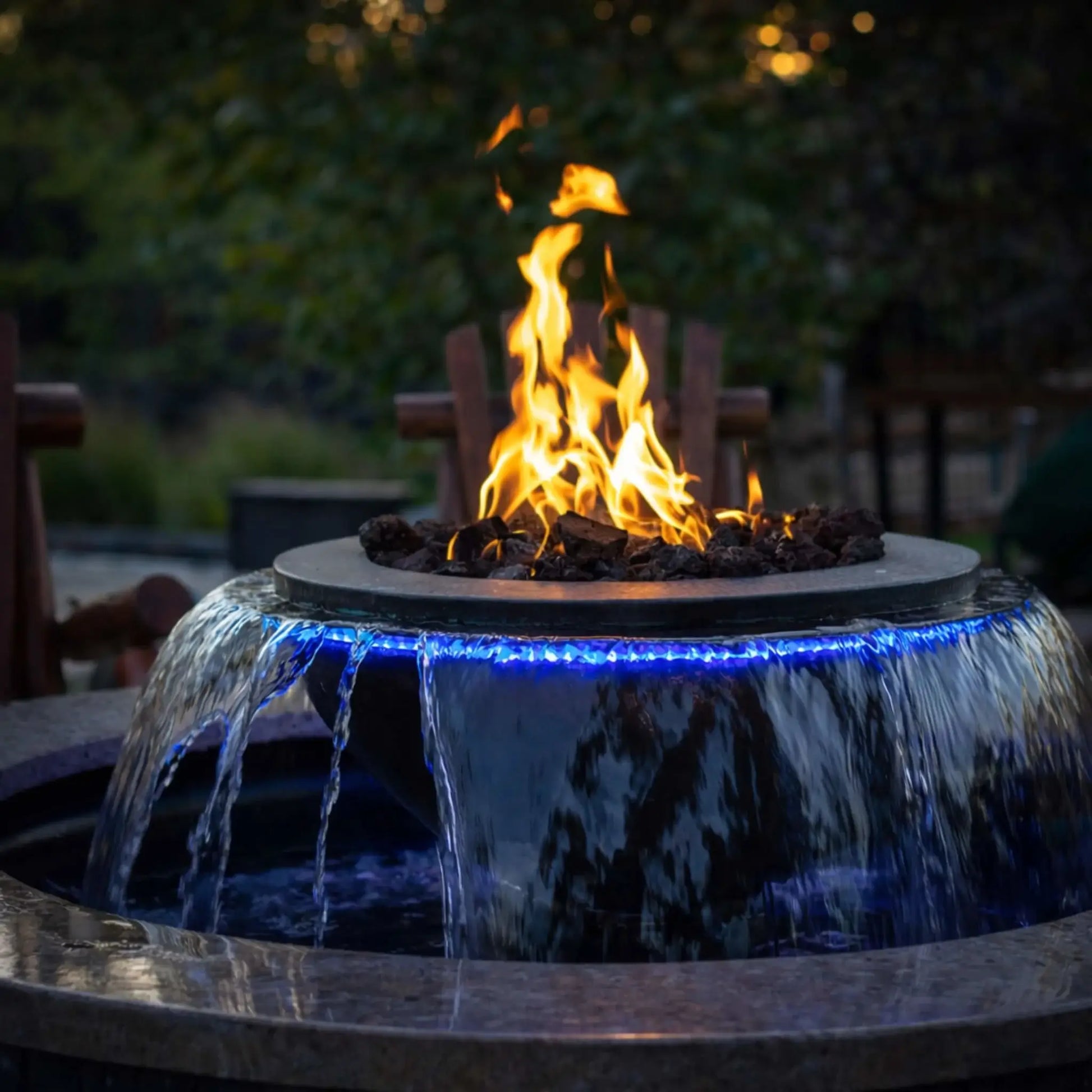 Modern fire and water feature combining a round fire bowl with cascading water, illuminated by blue LED lighting, with flames rising above lava rock while water spills over the edges into a circular basin in an evening outdoor setting.