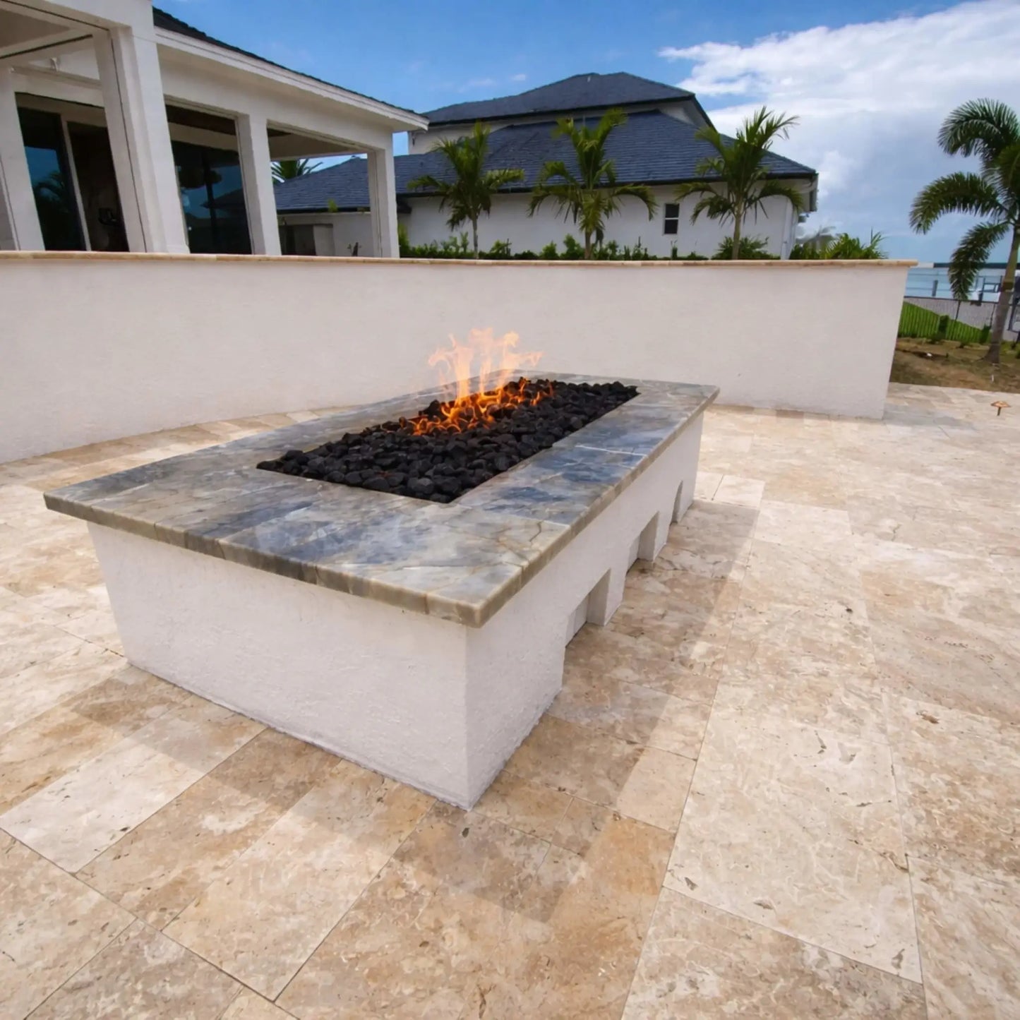 Modern rectangular gas fire pit table with a gray stone top and white base, filled with black lava rock and flames, placed on a light travertine patio near a coastal-style home with palm trees and a water view.