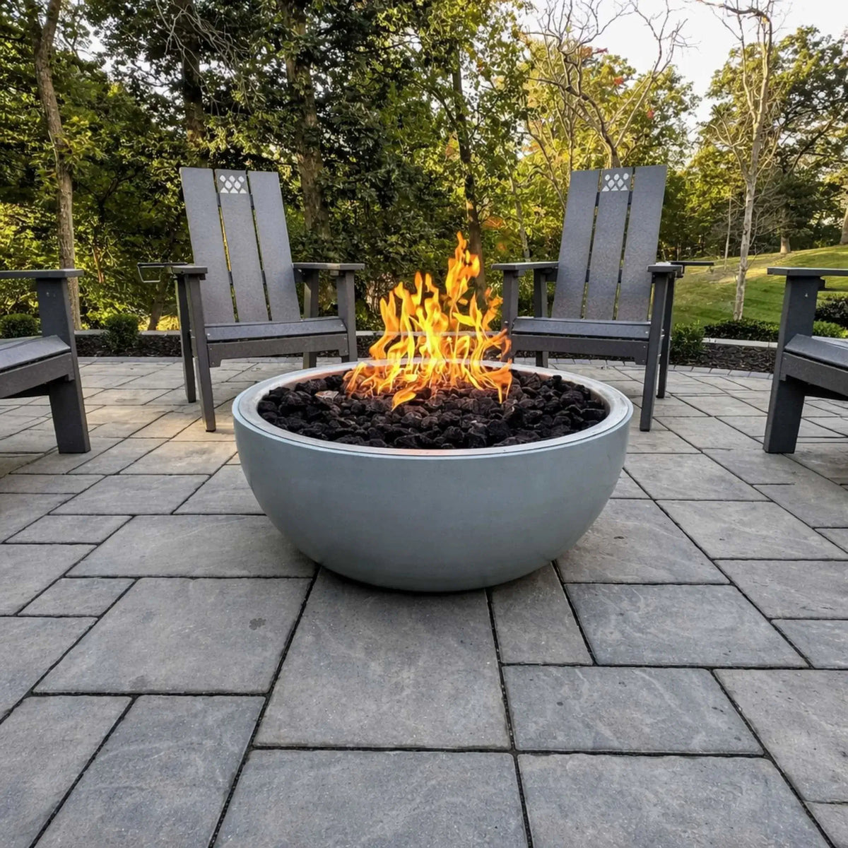 Round concrete fire bowl filled with black lava rock and bright flames, centered on a paved patio surrounded by Adirondack chairs and trees, creating a cozy backyard gathering space in a natural wooded setting.