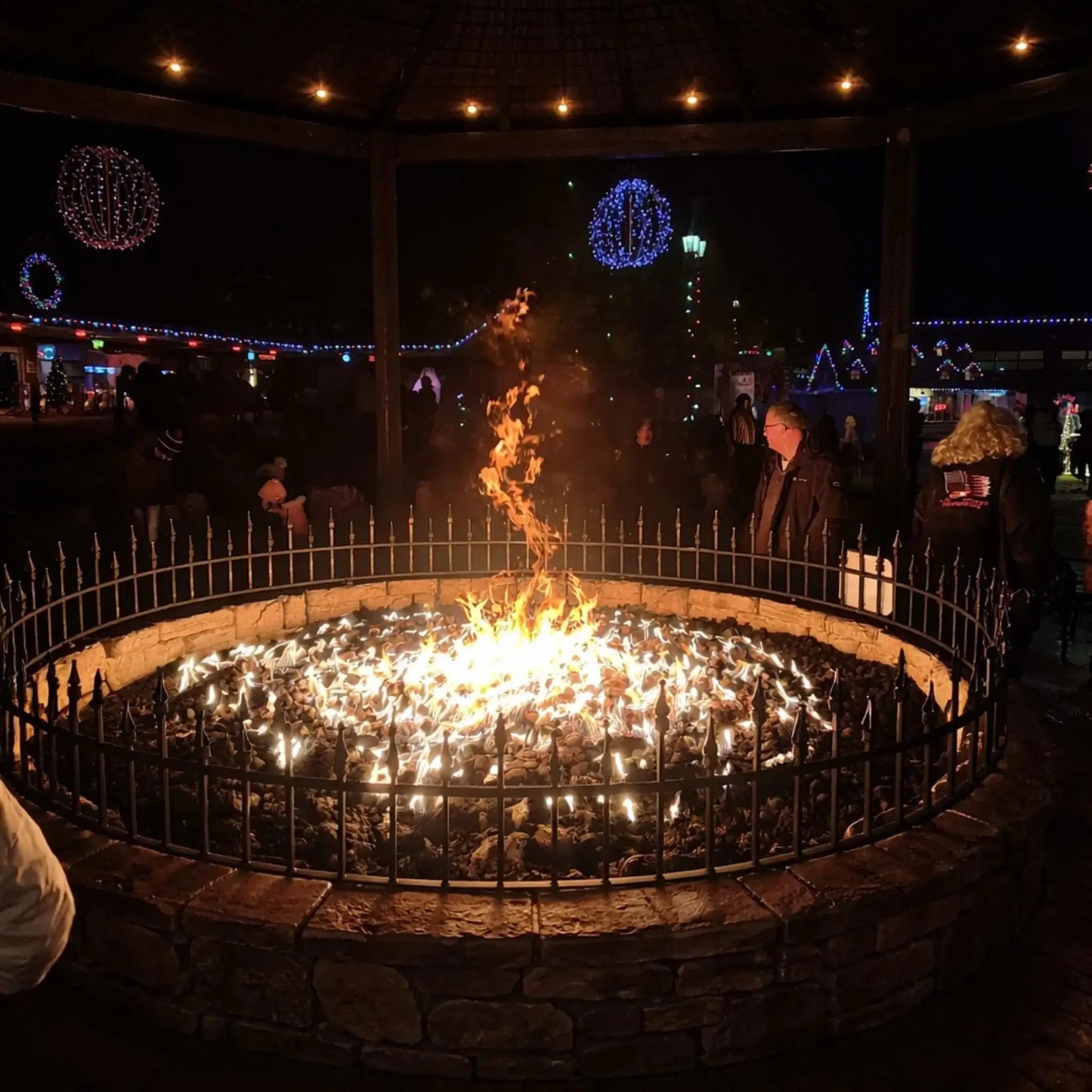 Large circular public fire pit filled with lava rock and surrounded by a metal safety railing, with a tall central flame feature glowing at night under a pavilion decorated with colorful holiday lights and people gathered around.