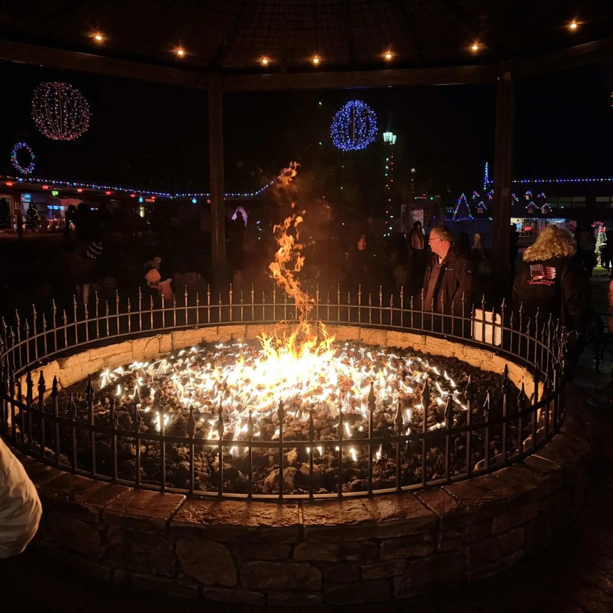 Large circular public fire pit filled with lava rock and surrounded by a metal safety railing, with a tall central flame feature glowing at night under a pavilion decorated with colorful holiday lights and people gathered around.