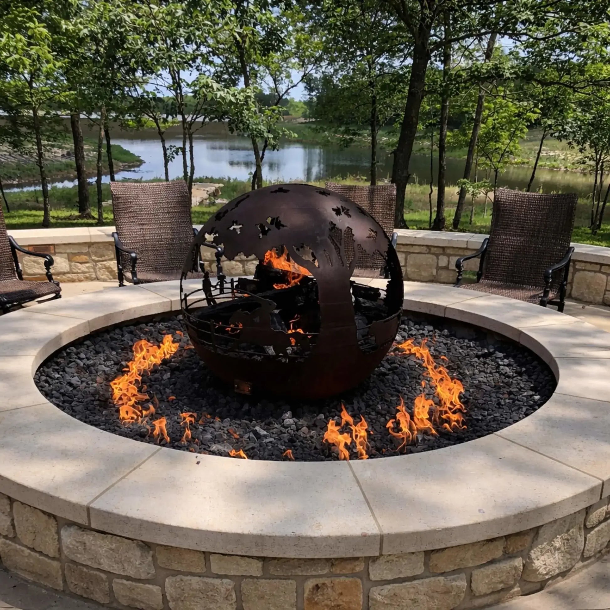 Custom circular fire pit with a stone surround and black lava rock, featuring a decorative metal globe sculpture centerpiece with flames rising around it, set in a scenic outdoor seating area overlooking a lake and trees.