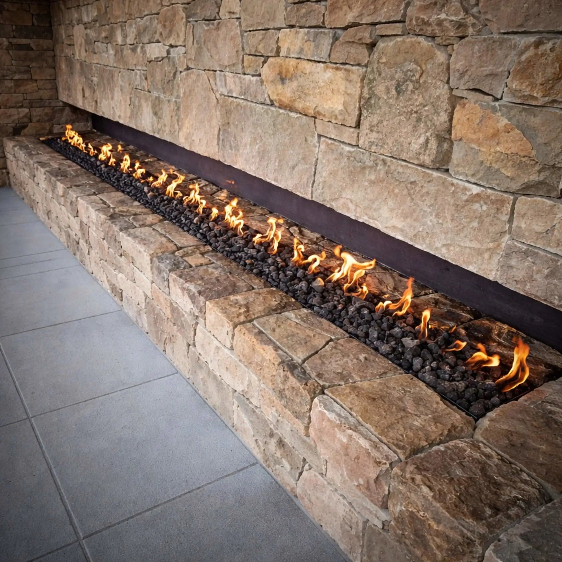 Long linear gas fire feature recessed into a stacked stone wall, filled with black lava rock and producing an even line of flames, integrated into a modern outdoor patio design with clean stonework and neutral tones.