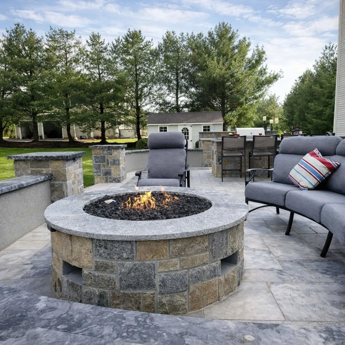 Round stone gas fire pit with a polished granite top and lava rock burner, surrounded by cushioned outdoor seating and a backyard patio with stone columns, bar area, and trees in the background for a relaxed residential setting.