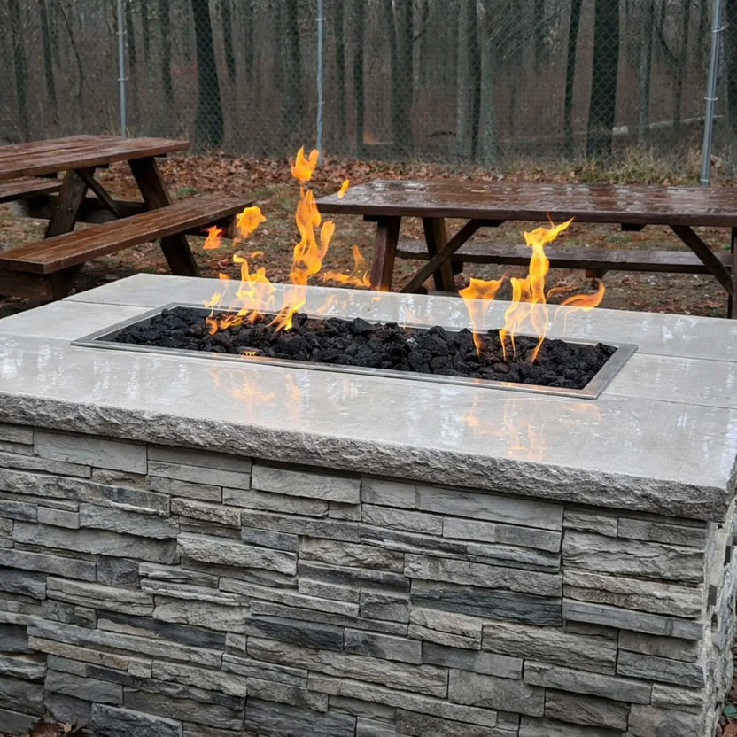 Rectangular outdoor fire table with a smooth light stone surface and inset burner tray filled with black lava rock, producing tall flames, positioned on a patio near wooden picnic tables and a fenced wooded area.