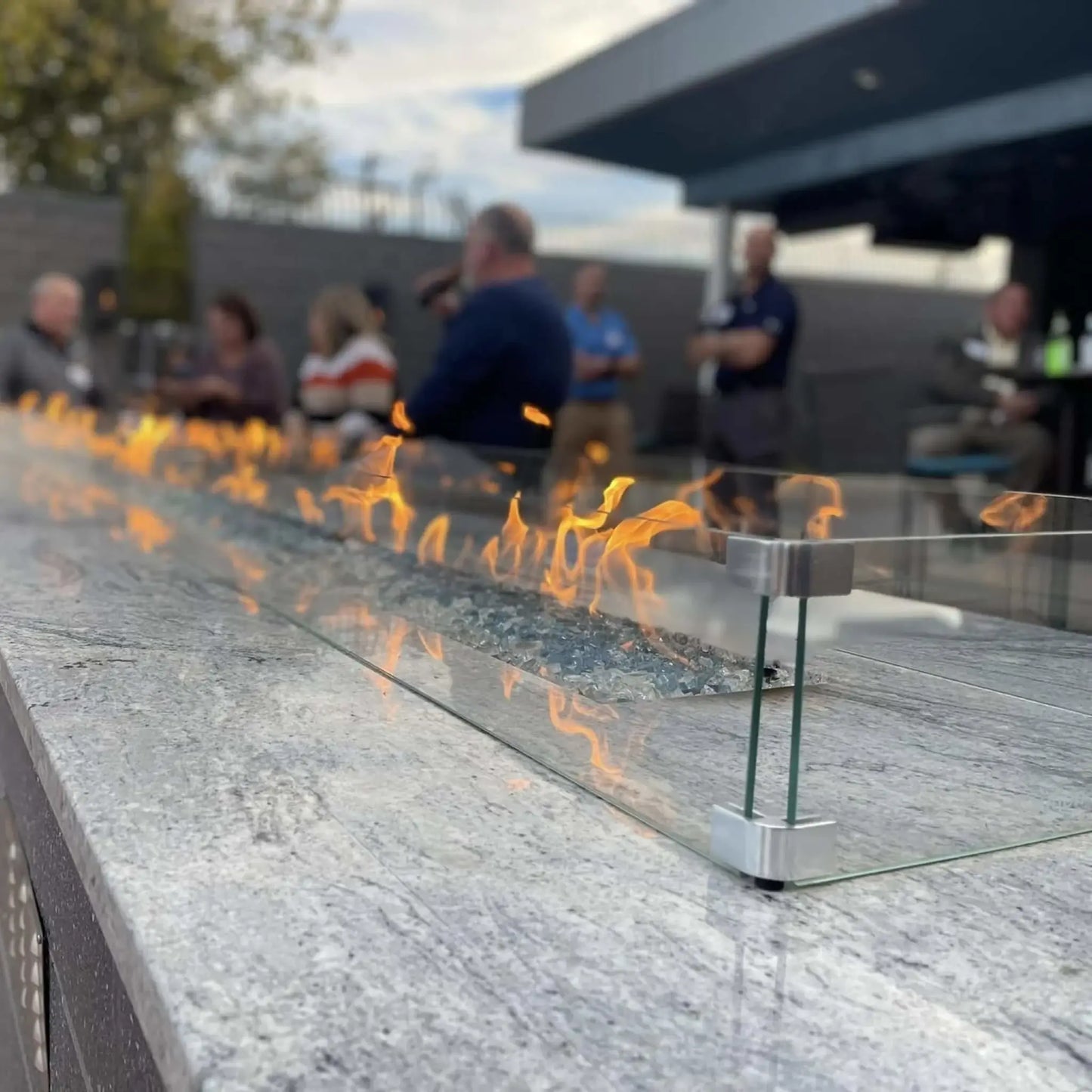 Close-up of a modern linear gas fire pit with blue fire glass and a clear tempered glass wind guard, mounted on a polished stone surface. Bright orange flames dance along the burner while a group of people gather in the softly blurred background, creating a warm outdoor social setting.