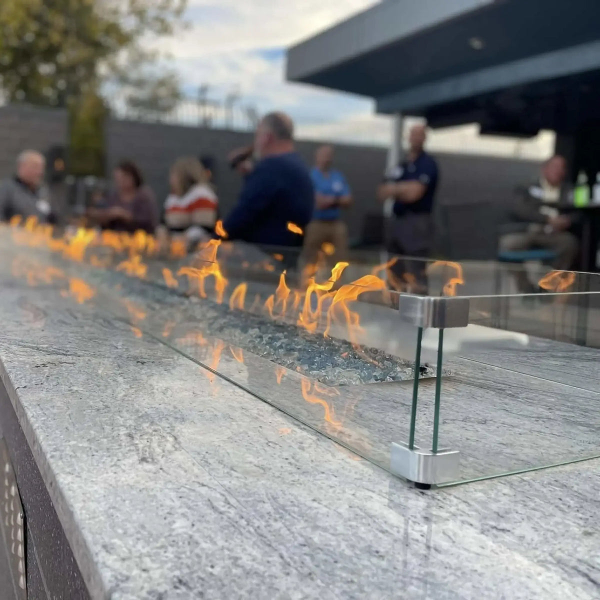 Close-up of a modern linear gas fire pit with blue fire glass and a clear tempered glass wind guard, mounted on a polished stone surface. Bright orange flames dance along the burner while a group of people gather in the softly blurred background, creating a warm outdoor social setting.