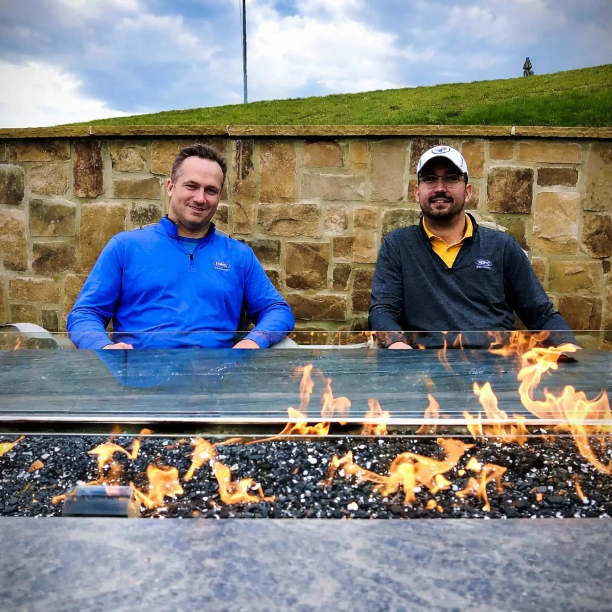 Two men sit behind a sleek linear gas fire pit filled with black fire glass, enclosed by a clear tempered glass wind guard. The flames stretch across the burner while a stone wall and grassy hillside create a relaxed outdoor setting.