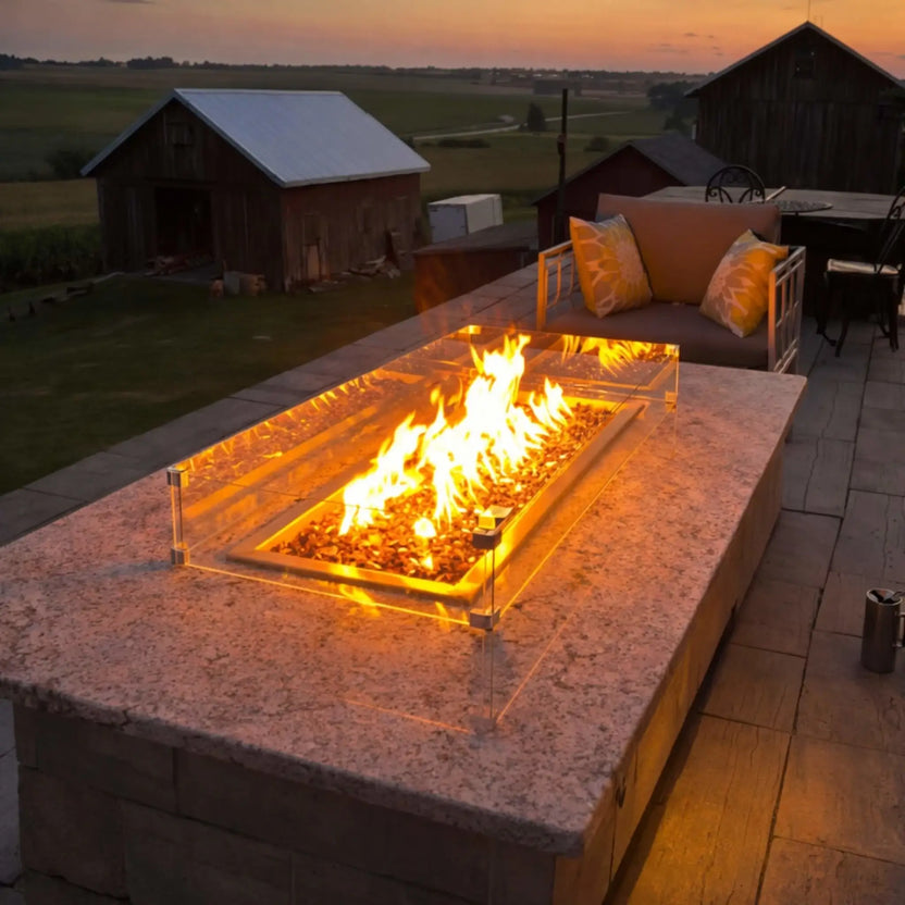 Rectangular outdoor fire pit table glowing at sunset, featuring a tempered glass wind guard and warm amber flames over fire glass. Rustic barns and open farmland in the background create a cozy, countryside evening atmosphere.