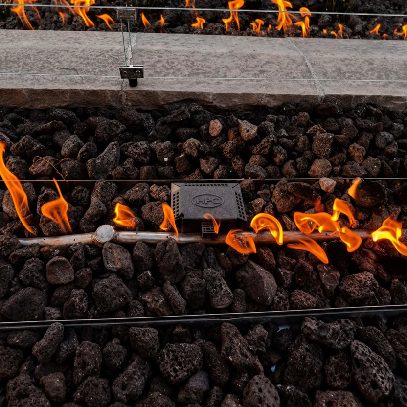 Close-up of a gas fire pit burner surrounded by black lava rocks, with bright flames rising around a metal burner tube and ignition component. The tempered glass wind guard sits just above, adding a clean, modern finish.