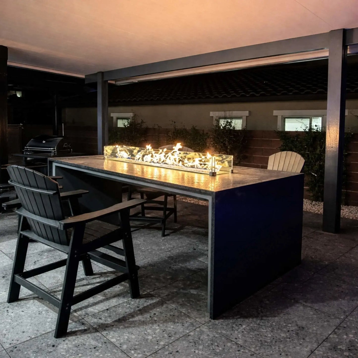 Modern outdoor patio dining area featuring a long rectangular fire table with a glass wind guard, glowing warmly at night. Adirondack chairs and a grill sit nearby under a covered structure, creating a comfortable backyard entertaining space.