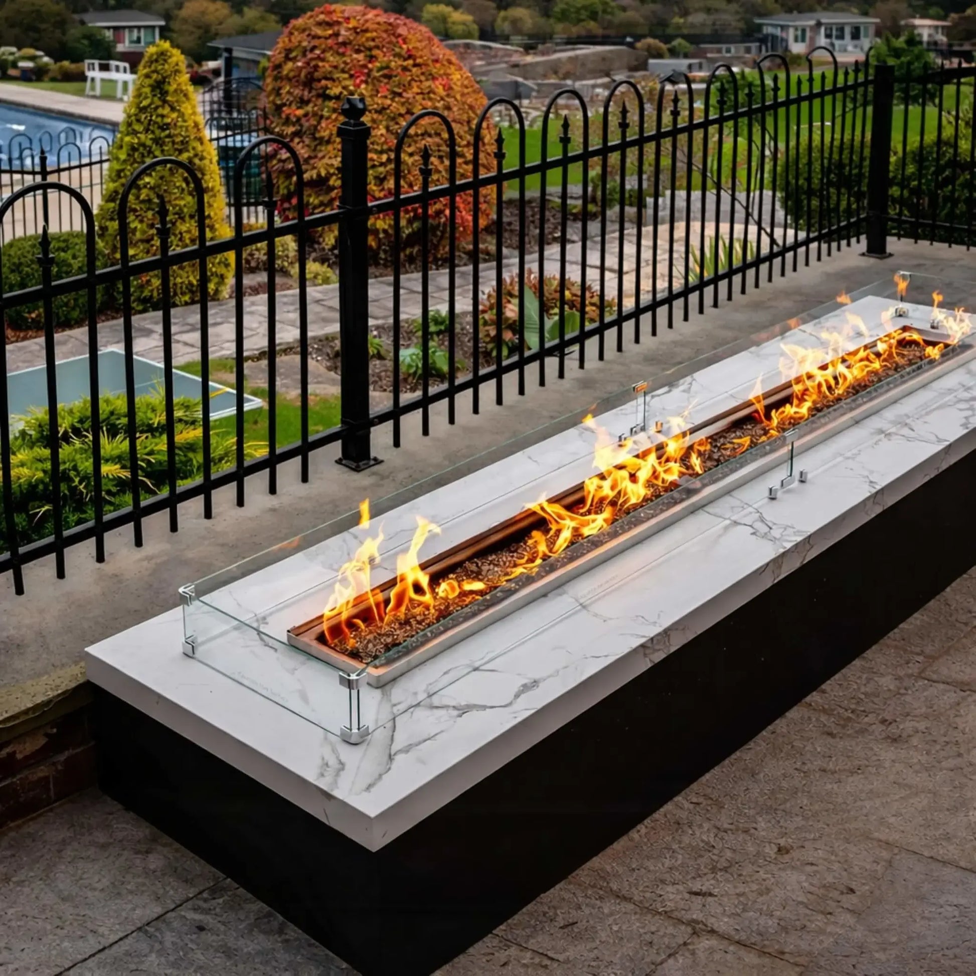 Linear fire pit table with a marble-style top and glass wind guard, installed along a patio edge overlooking landscaped grounds. Black metal railing frames the space while bright flames run the full length of the burner.