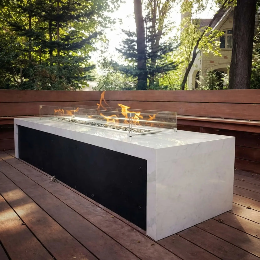 Contemporary rectangular fire pit table with a white stone top and glass wind guard, set on a wooden deck surrounded by trees. Soft flames rise above decorative fire media, creating a peaceful backyard retreat.