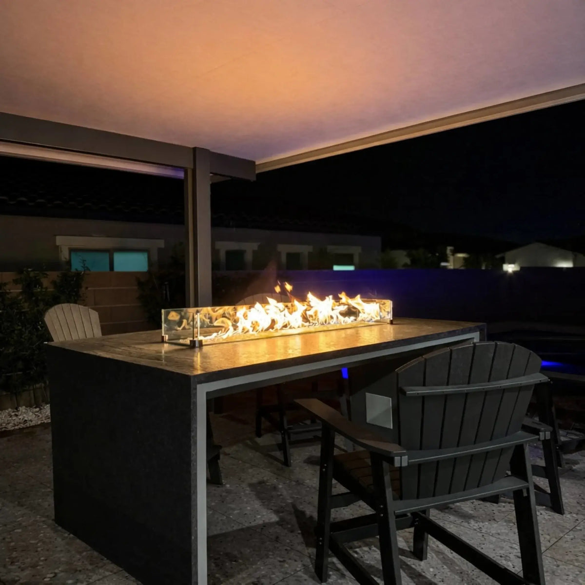 Outdoor fire table glowing at night beneath a covered patio, featuring a long linear burner with a tempered glass wind guard. Adirondack chairs surround the table, and subtle lighting highlights the modern backyard setting.