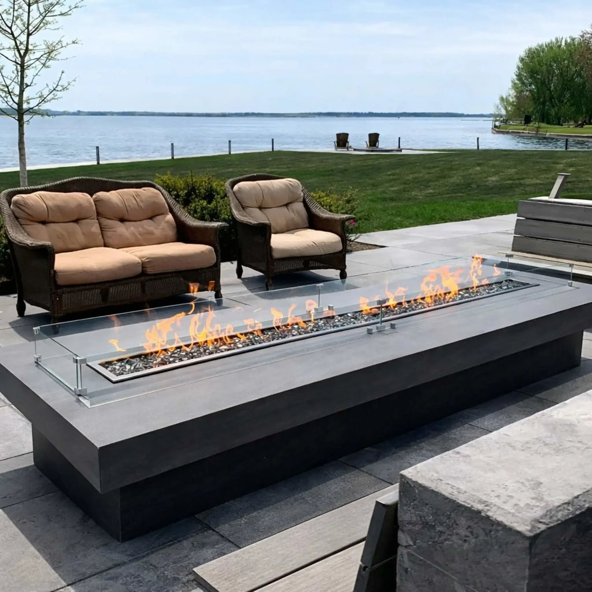 Large rectangular fire pit table with a sleek linear flame and glass wind guard, positioned on a patio overlooking a lake. Cushioned outdoor seating and a wide open waterfront view create a high-end outdoor living space.