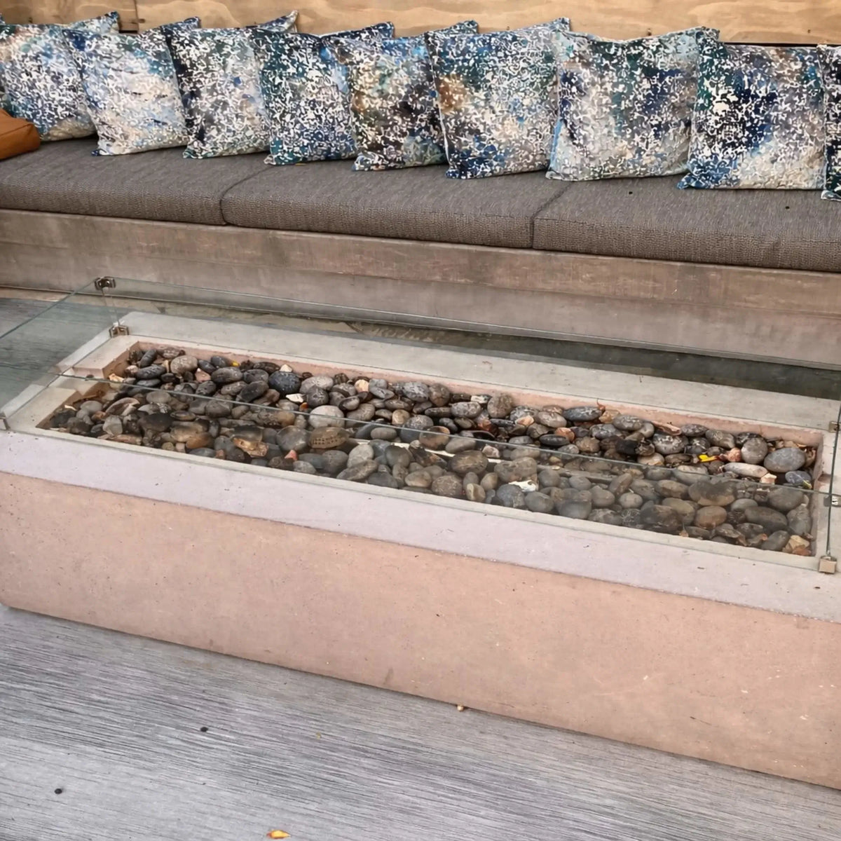 Unlit linear fire pit filled with smooth river stones and enclosed by a clear glass wind guard, set in front of a cushioned outdoor bench with patterned pillows. The scene highlights the clean design and materials before ignition.