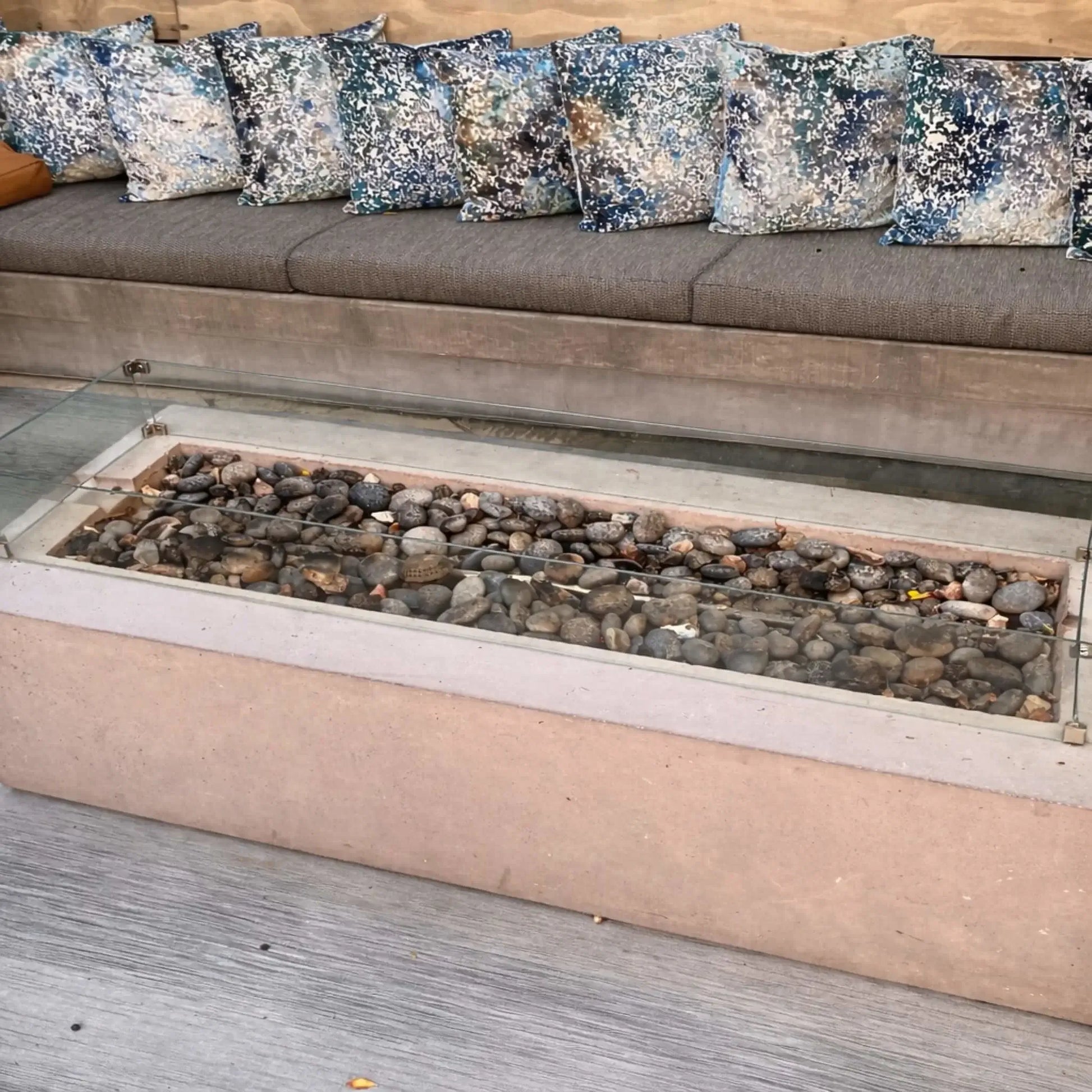 Unlit linear fire pit filled with smooth river stones and enclosed by a clear glass wind guard, set in front of a cushioned outdoor bench with patterned pillows. The scene highlights the clean design and materials before ignition.