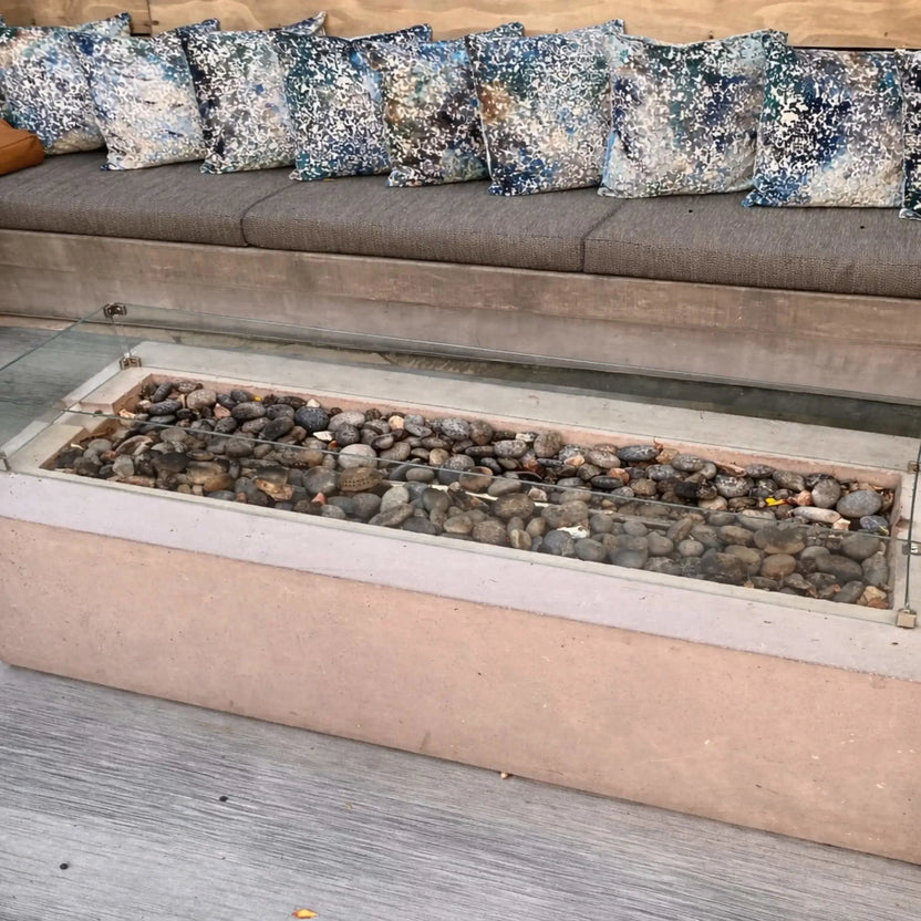 Unlit linear fire pit filled with smooth river stones and enclosed by a clear glass wind guard, set in front of a cushioned outdoor bench with patterned pillows. The scene highlights the clean design and materials before ignition.