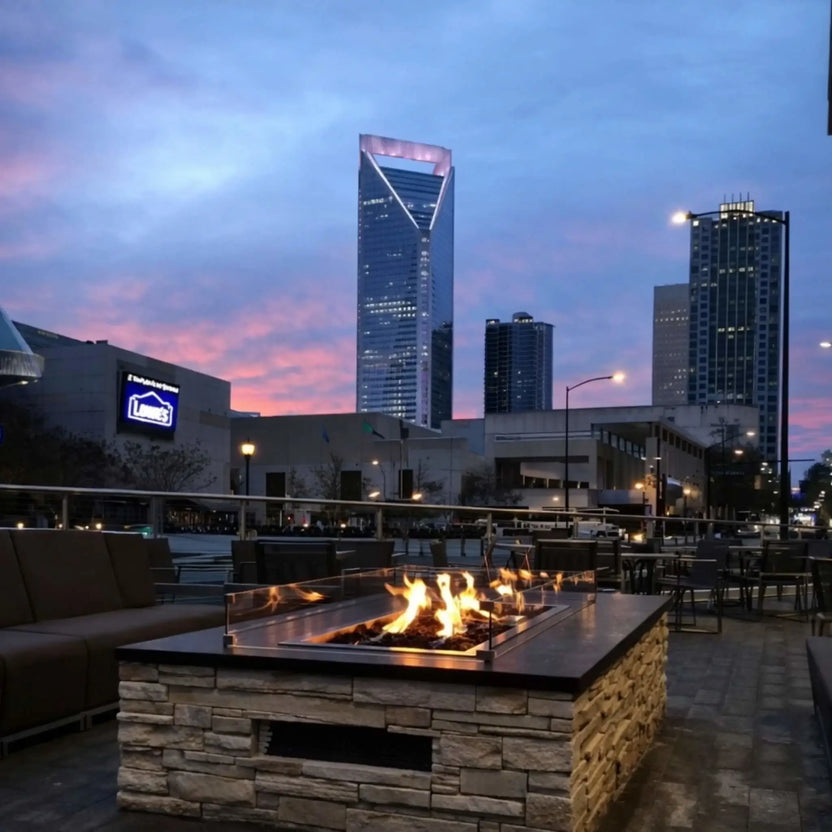 Modern outdoor lounge patio with a rectangular gas fire pit table featuring a tempered glass wind guard and natural stone base. Flames glow against a dramatic city skyline at sunset, creating an upscale urban outdoor living space.