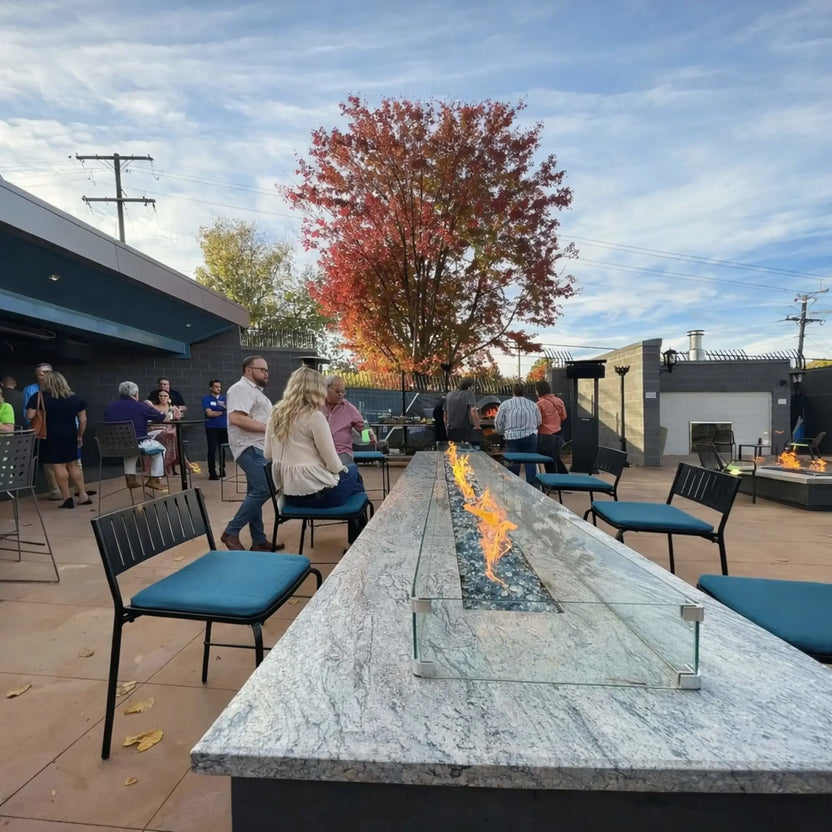 Wide view of a long rectangular fire pit table with a glass wind guard and blue fire glass centerpiece, surrounded by patio chairs and guests mingling outdoors. A vibrant autumn tree adds color to the scene, highlighting a stylish commercial outdoor lounge environment.