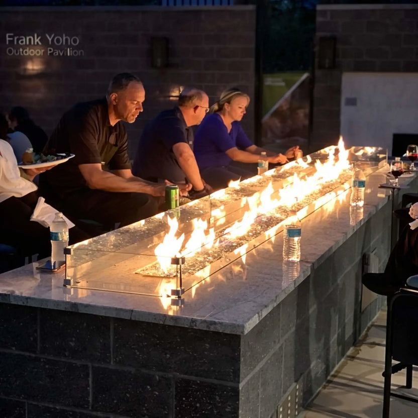 Group of people seated around a long linear fire pit table with a glass wind guard at an outdoor pavilion. Bright flames run the length of the burner while drinks and conversation create a warm, social gathering atmosphere.
