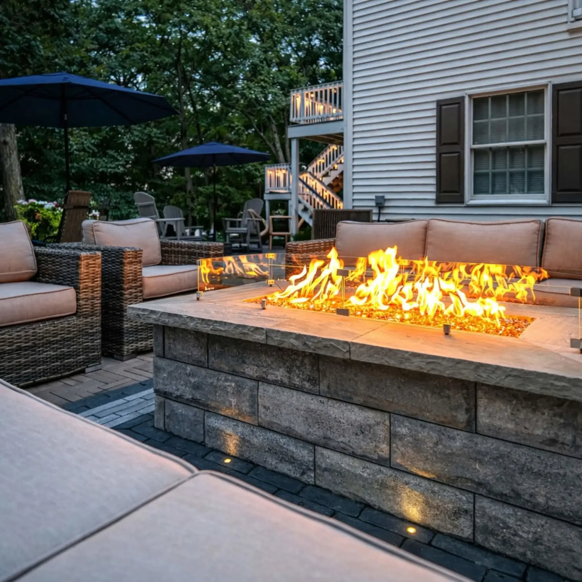 Backyard patio with woven outdoor seating surrounding a rectangular fire pit table topped with a glass wind guard. Vibrant flames rise over fire glass while a multi-level deck and trees create a cozy residential outdoor setting.