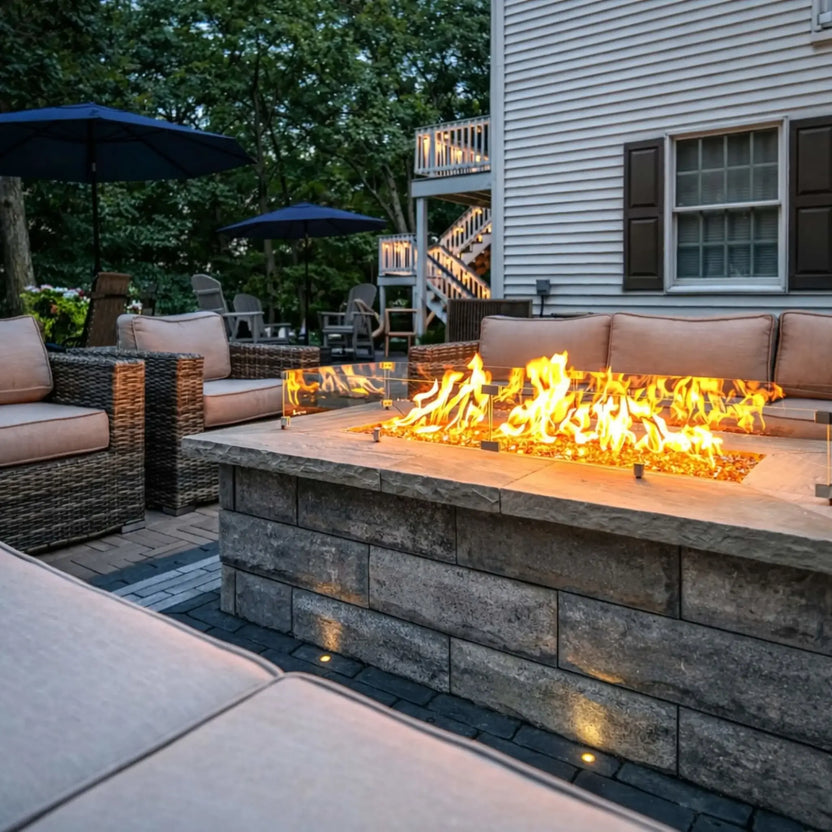 Backyard patio with woven outdoor seating surrounding a rectangular fire pit table topped with a glass wind guard. Vibrant flames rise over fire glass while a multi-level deck and trees create a cozy residential outdoor setting.