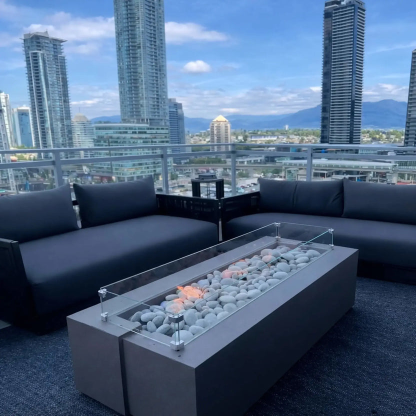 Rooftop patio with modern black seating and a linear fire pit table filled with smooth stones and enclosed by a glass wind guard. City high-rise buildings and distant mountains provide a scenic urban skyline backdrop.