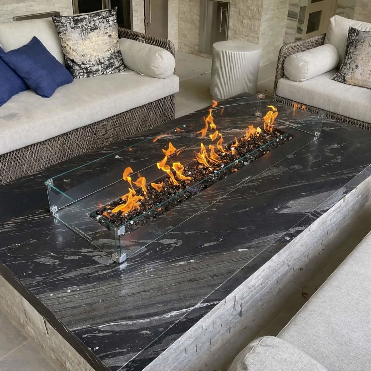 Contemporary outdoor seating area featuring a black marble-style fire table with a linear burner and tempered glass wind guard. Flames dance above dark fire glass while cushioned sofas and decorative pillows add a luxury feel.