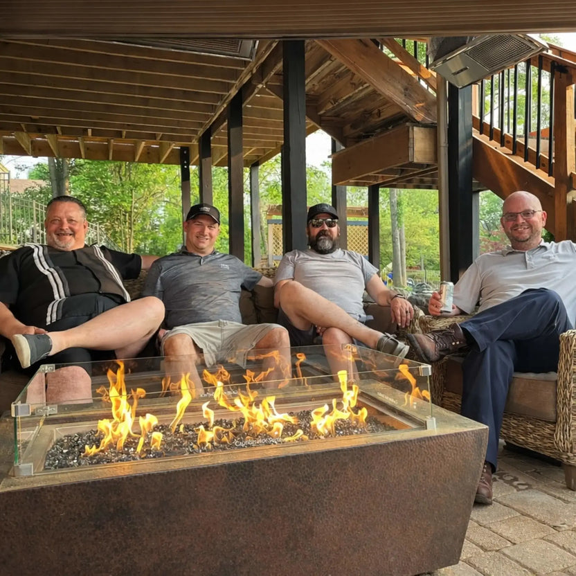 Four men relaxing on a covered patio around a rectangular fire pit table with a glass wind guard. Flames stretch across black fire glass, creating a warm focal point in a casual outdoor gathering space with wood beams overhead.