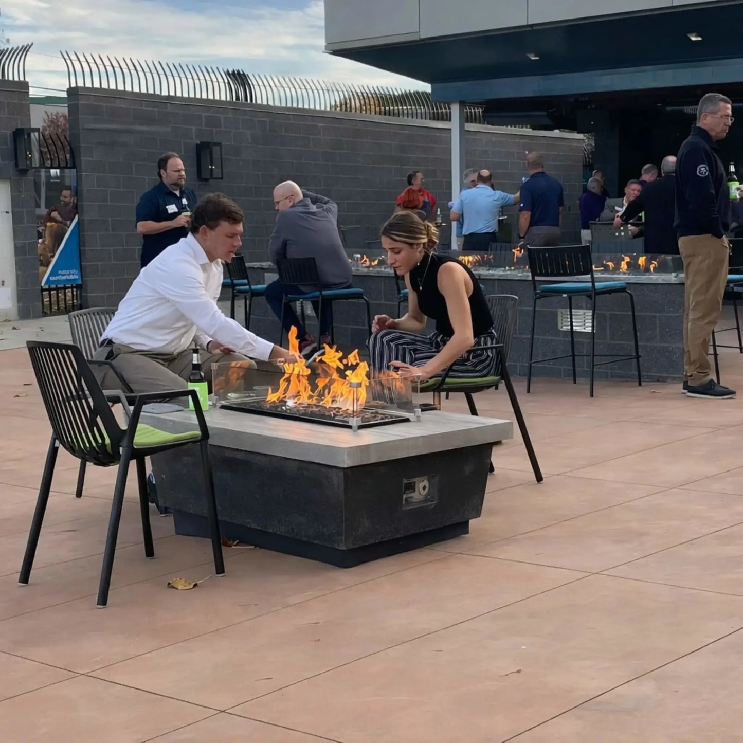 Two people seated at a square gas fire pit table with a tempered glass wind guard, adjusting the flames over decorative fire media. The setting features a modern outdoor patio with bar seating and guests socializing in the background.