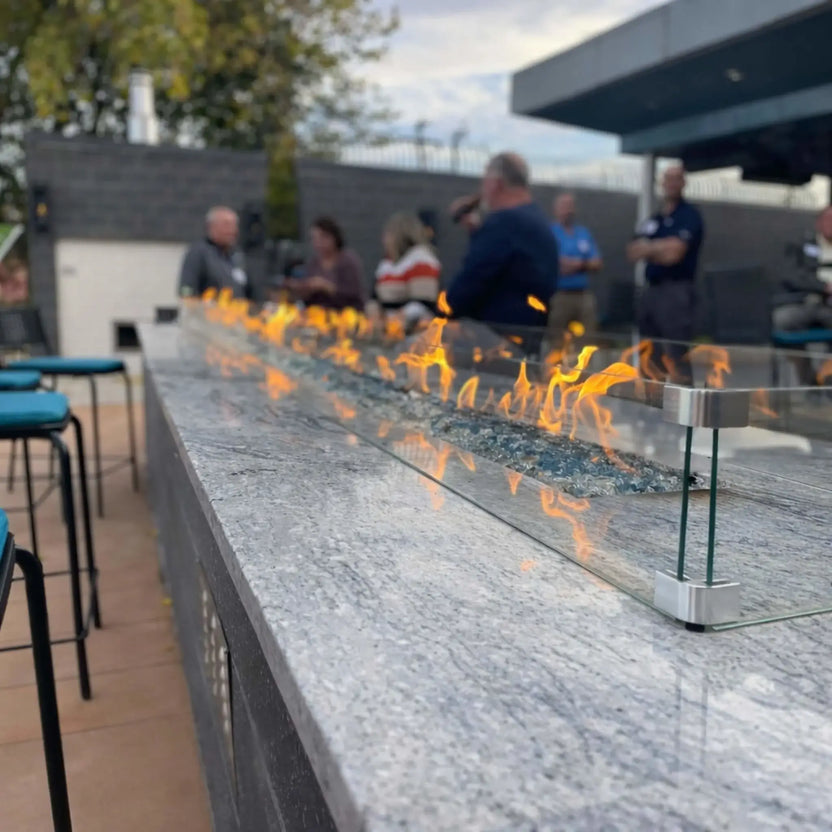 Angled close-up of a sleek linear fire pit burner filled with blue fire glass and enclosed by a clear glass wind guard. The flames reflect across the polished stone surface while a group of people stands in the softly out-of-focus background.