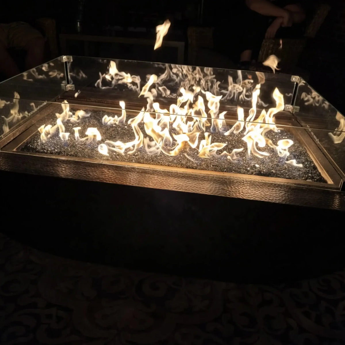 Evening scene of a rectangular fire pit filled with dark fire glass, surrounded by a clear tempered glass wind guard. Bright flames flicker vividly against a dark background, creating a dramatic and cozy outdoor ambiance.