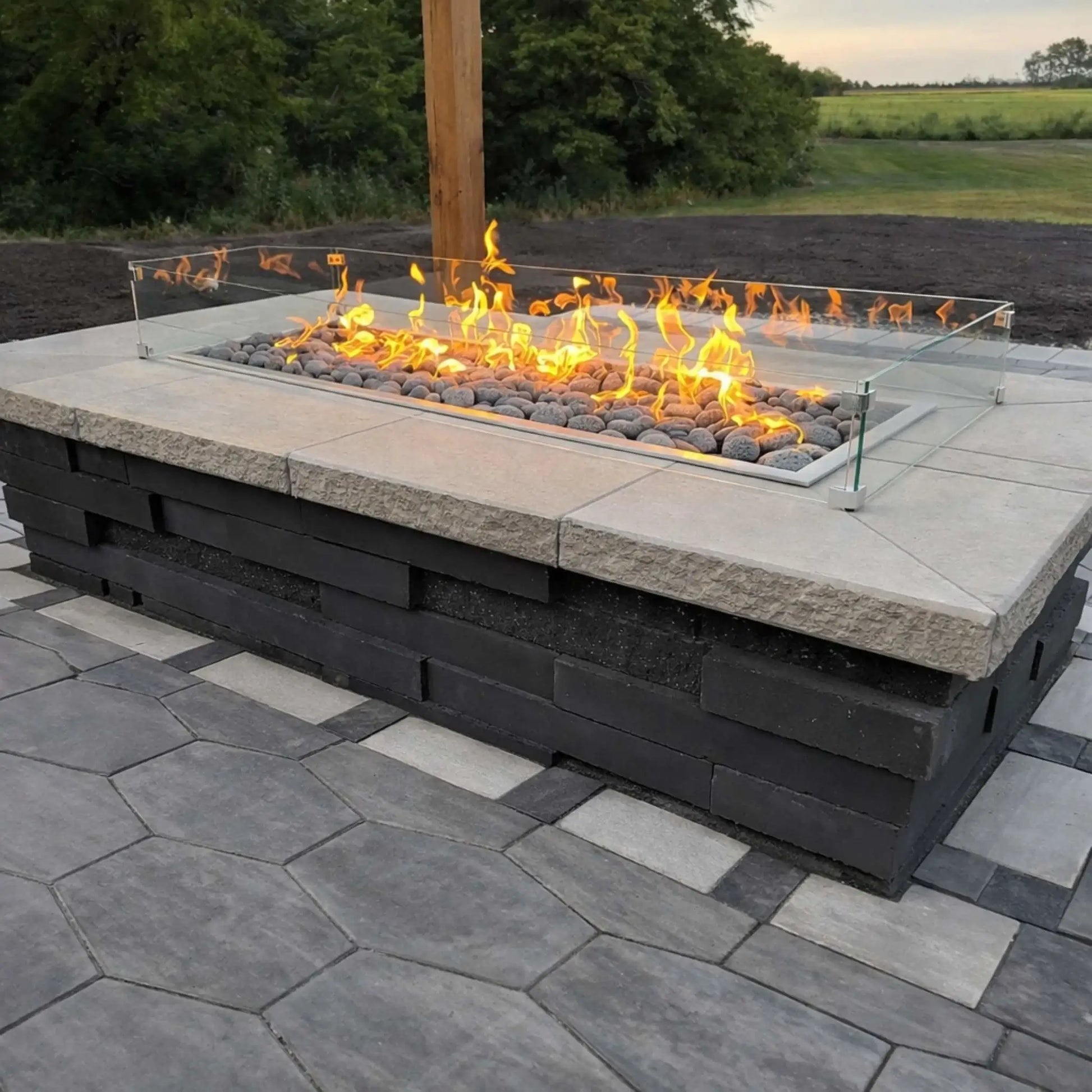 Outdoor patio fire feature with a long linear burner filled with smooth gray stones and protected by a glass wind guard. The fire pit is built into a block-style base and sits on a patterned paver surface, overlooking a natural landscape.