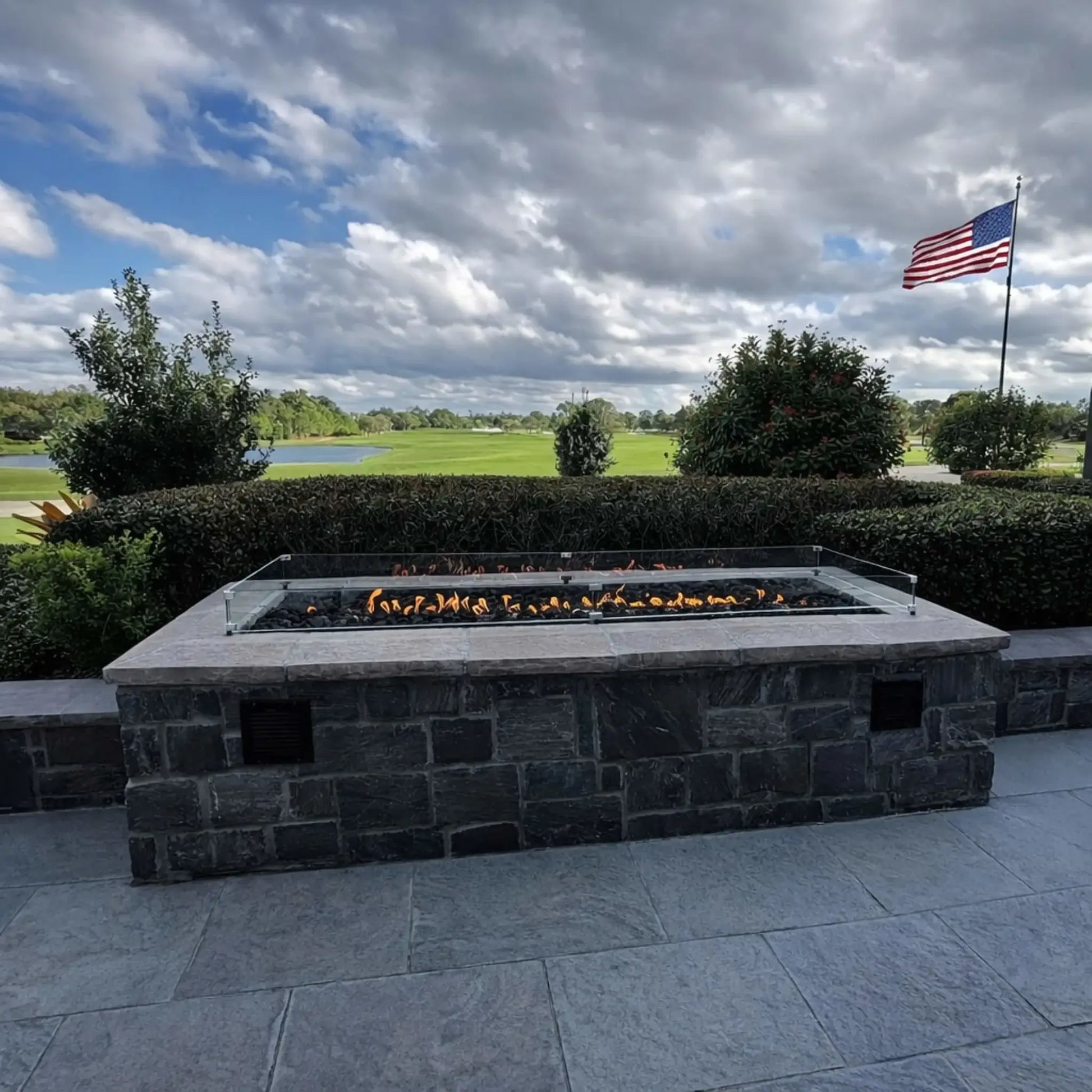 Large rectangular stone fire pit with a glass wind guard installed on a landscaped patio, overlooking a golf course and water feature. An American flag waves in the background under a cloudy sky, adding a scenic, upscale outdoor setting.