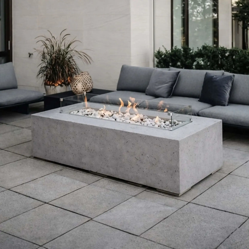 Modern concrete fire pit table with white decorative stones and a tempered glass wind guard, positioned in a contemporary outdoor lounge with gray cushioned seating and paver flooring. Soft flames create a relaxing, upscale patio atmosphere.