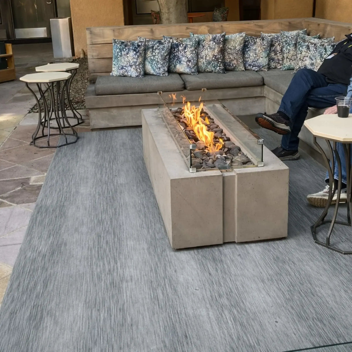 Rectangular concrete fire pit with dark lava rocks and a glass wind guard, placed in a cozy outdoor seating area with sectional furniture and patterned cushions. A person relaxes nearby, highlighting a comfortable residential patio setting.