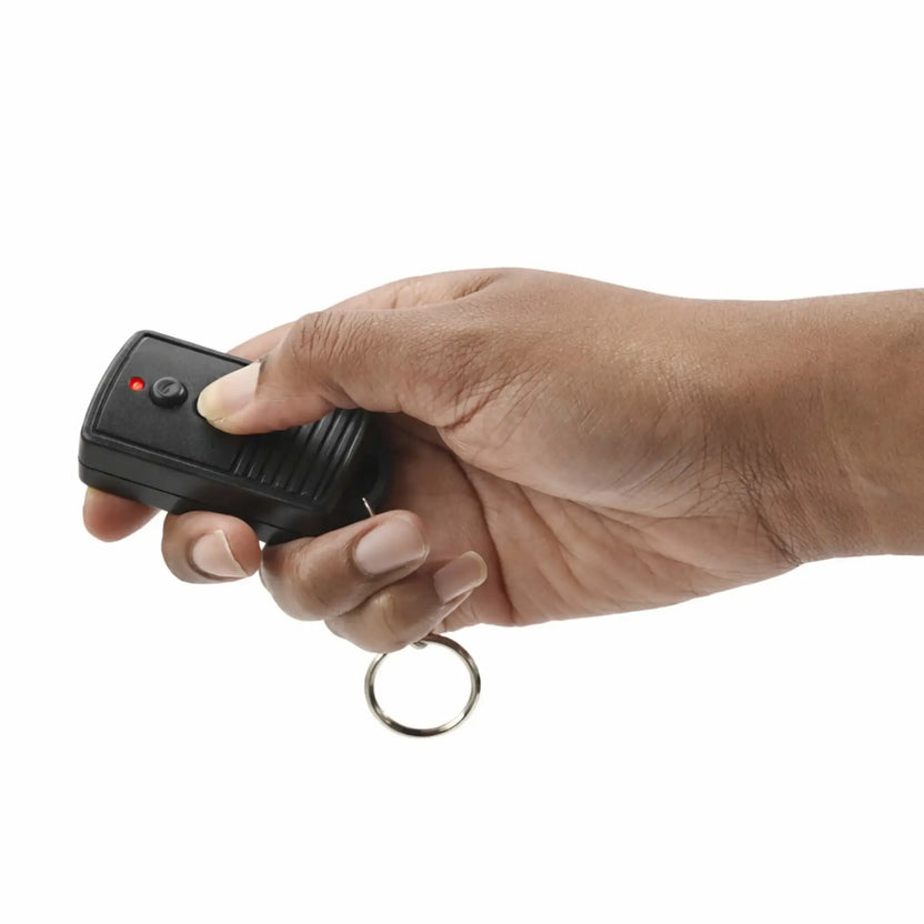 Lifestyle image showing a person holding a black handheld on/off remote control with a keyring attachment, actively pressing one of the buttons. The remote features a red LED indicator light and textured grip, demonstrating ease of use and portability for controlling a fire pit or fireplace system.