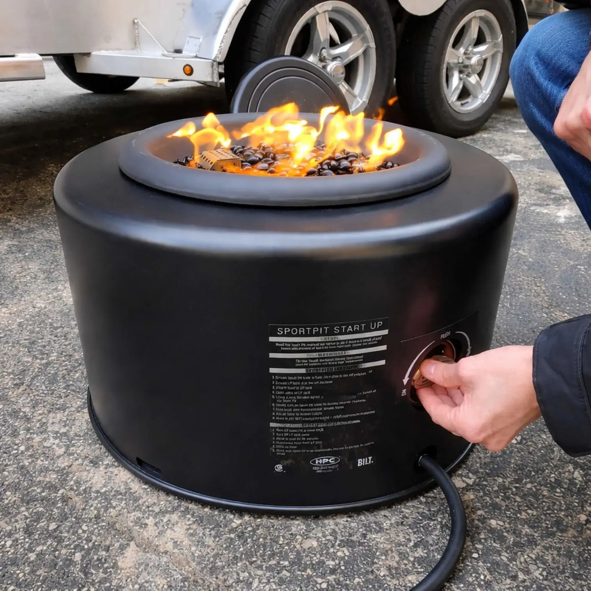 Close-up of the HPC Portable Sport Pit tailgate fire pit in use, showing a hand turning the control knob to ignite flames over black lava rock. The compact round fire pit sits on pavement near a trailer, highlighting its portable propane setup and easy-start ignition system.