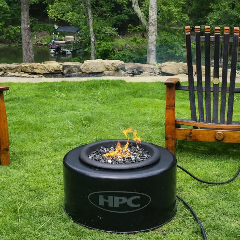 HPC Portable Sport Pit fire pit set on a grassy lakeside lawn, with flames burning over lava rock. Surrounded by wooden chairs and a scenic pond with a docked boat in the background, this Portable Sport Pit tailgate fire setup highlights relaxing outdoor use in a peaceful backyard setting.