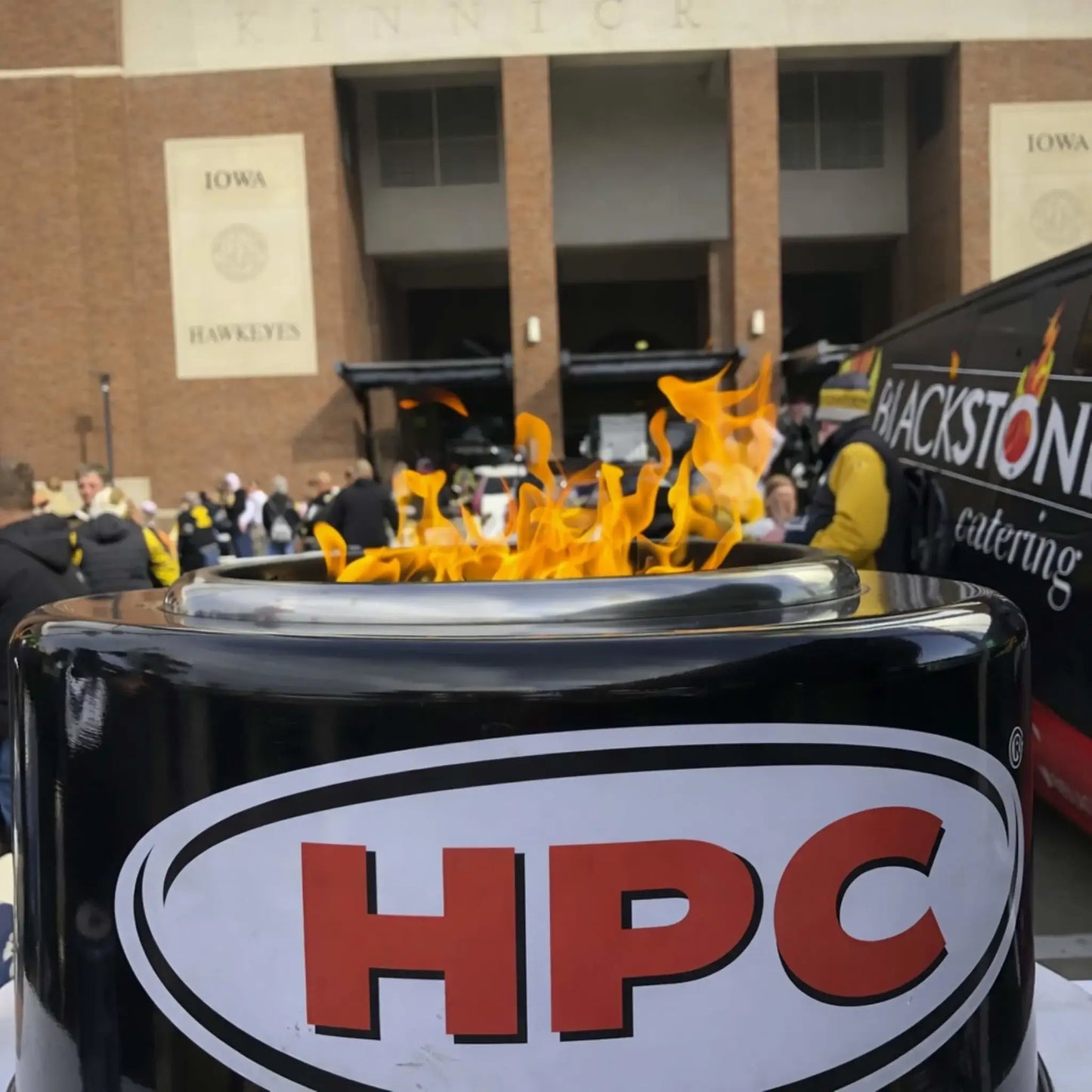 Close-up of the HPC Portable Sport Pit fire pit with bright flames in front of a stadium during a tailgating event. The HPC logo is clearly visible on the unit, with a crowd and vendor setup in the background, showcasing the Portable Sport Pit Tailgate Fire in a real game-day environment.