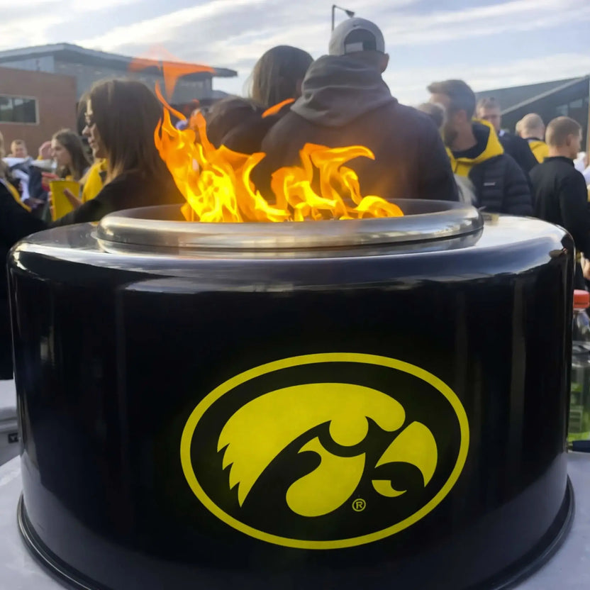 HPC Portable Sport Pit tailgate fire pit in action with Iowa Hawkeyes branding, surrounded by fans at an outdoor event. Flames rise from the top burner as people gather nearby, emphasizing the Portable Sport Pit’s role in social tailgating and outdoor entertainment settings.