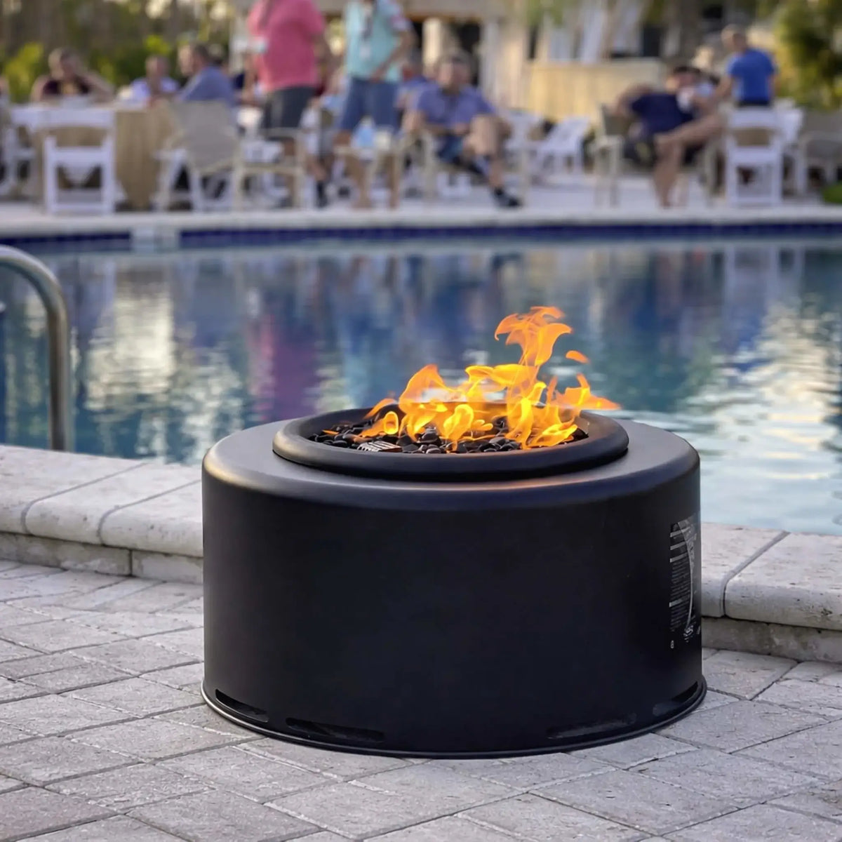 HPC Portable Sport Pit fire pit burning beside a pool, with bright flames rising from black lava rock. The round, low-profile design is shown in a social outdoor setting with people relaxing in the background, emphasizing its use for backyard gatherings and poolside ambiance.