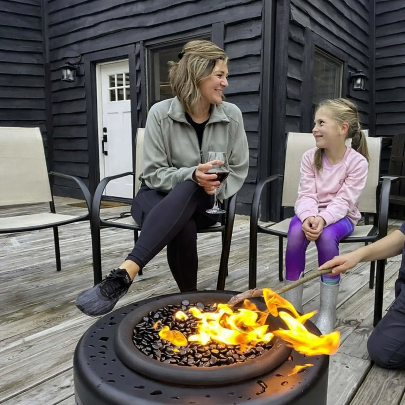 HPC Portable Sport Pit fire pit on a wooden deck, surrounded by a woman and child enjoying the warmth. Flames glow over lava rock while someone roasts a marshmallow, capturing a cozy family moment and showcasing the fire pit’s safe, portable outdoor use.