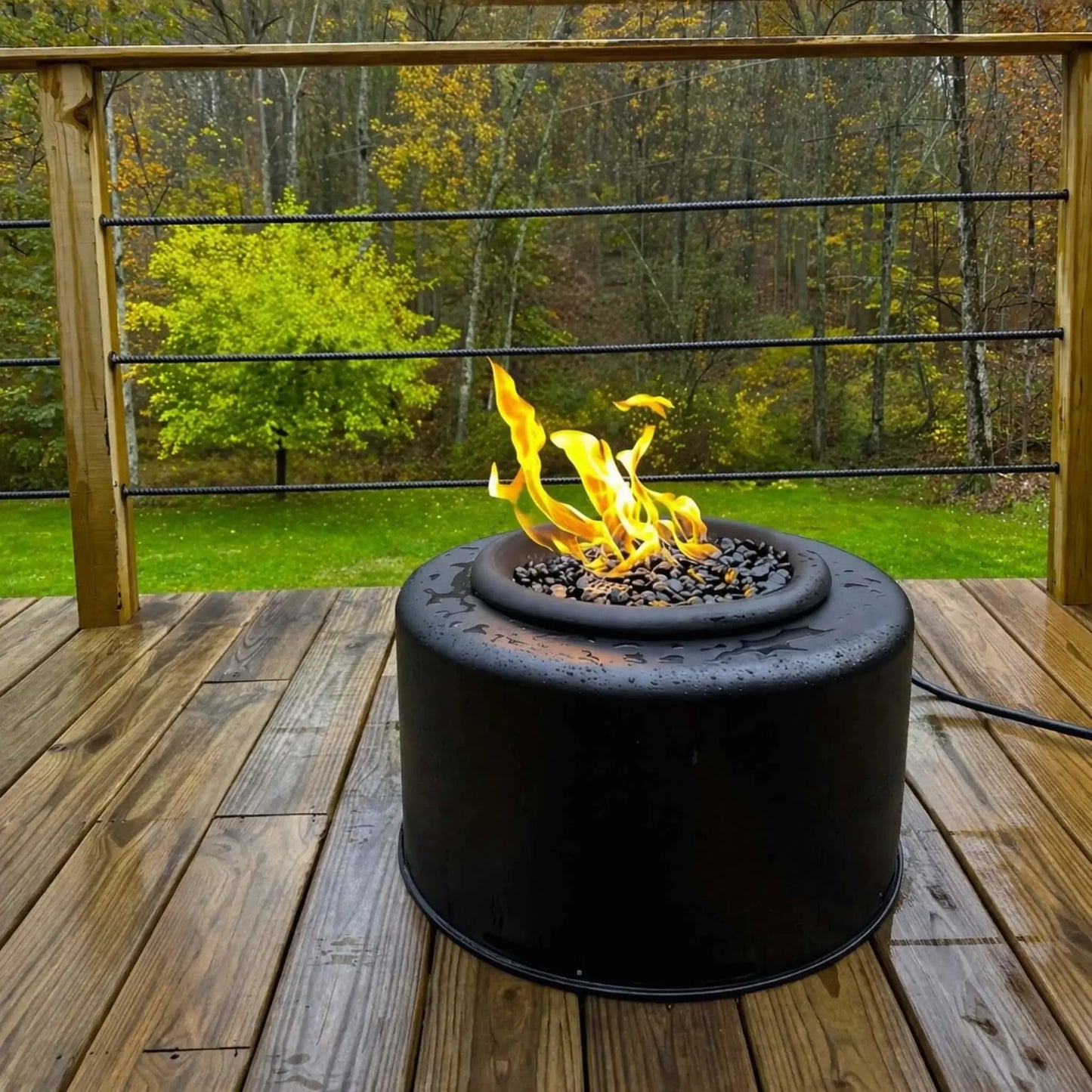 Portable Sport Pit fire pit by HPC placed on a wet wooden deck with a scenic wooded backyard. Flames flicker above black lava rock despite damp conditions, demonstrating durability and all-weather performance for outdoor fire features.