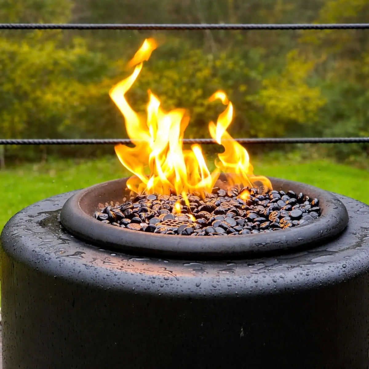 Tight close-up of the HPC Portable Sport Pit burner area with vivid yellow flames dancing over black lava rock. Water droplets on the fire pit surface highlight weather resistance, while the blurred green background adds a natural outdoor setting.