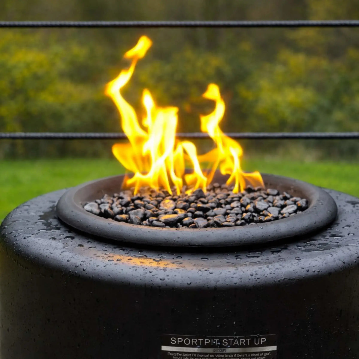 Another close-up angle of the HPC Portable Sport Pit fire pit, focusing on the flame pattern and lava rock media. The rounded edge and moisture on the surface emphasize durability and consistent flame performance in outdoor environments.